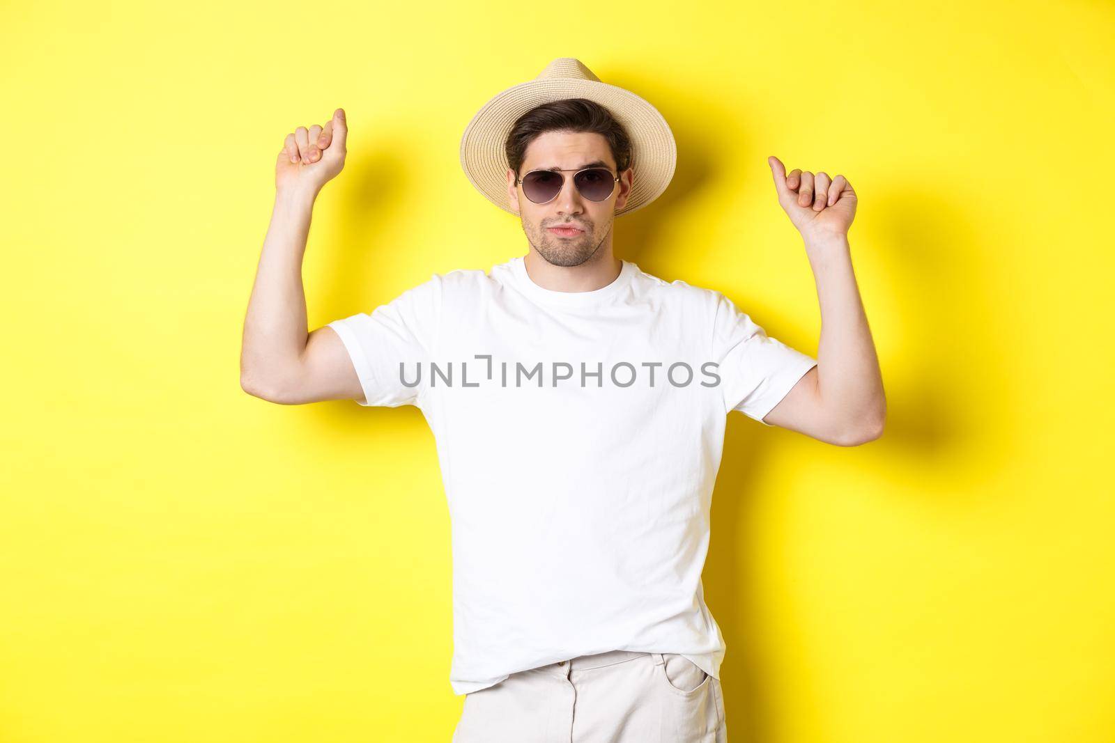 Tourism, travelling and holidays concept. Man tourist enjoying vacation, dancing in straw hat and sunglasses, posing against yellow background.