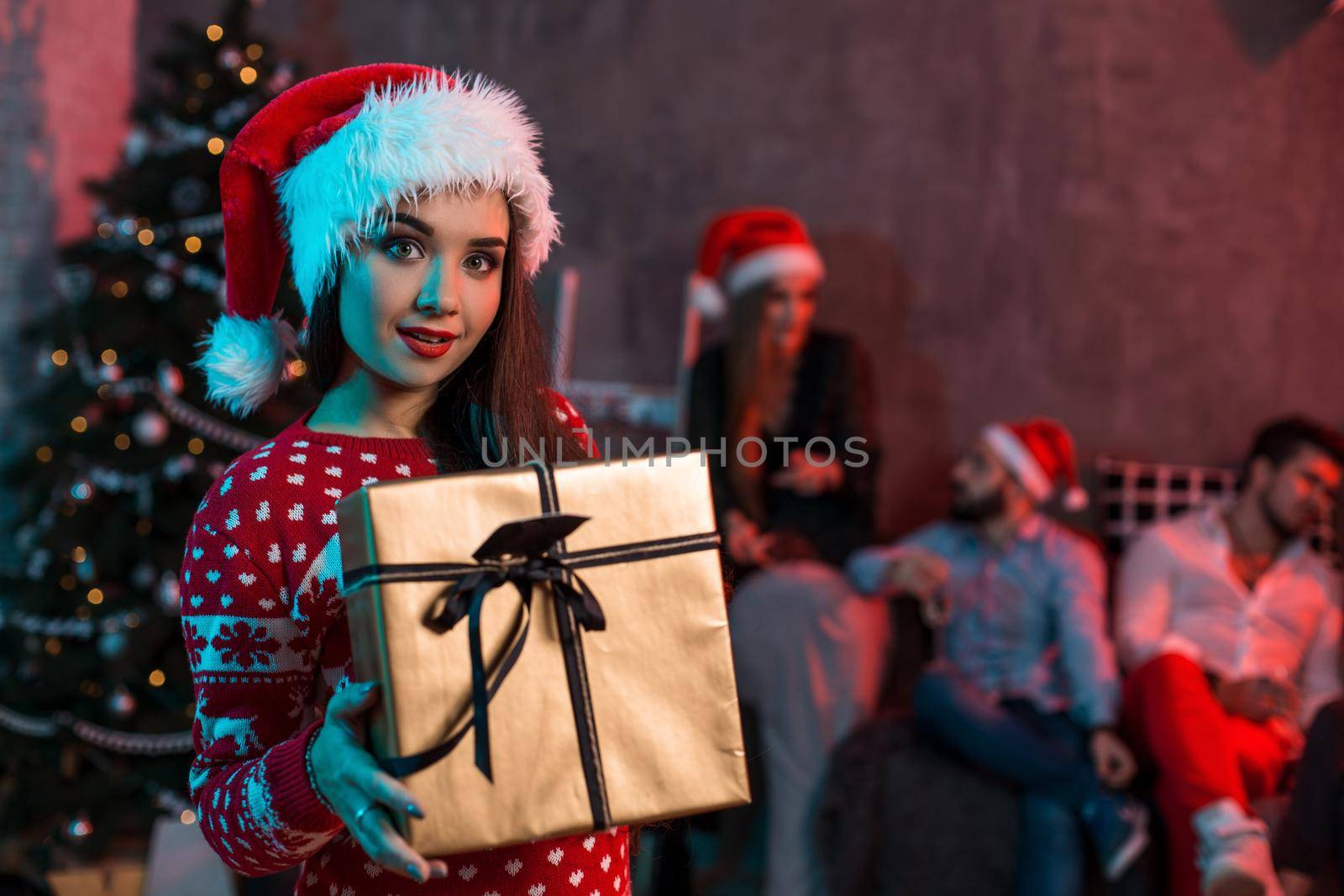 Portrait of a young woman with a gold giftbox at home on the foreground. Beautiful brunette in a Santa hat, red costume with deers by nazarovsergey
