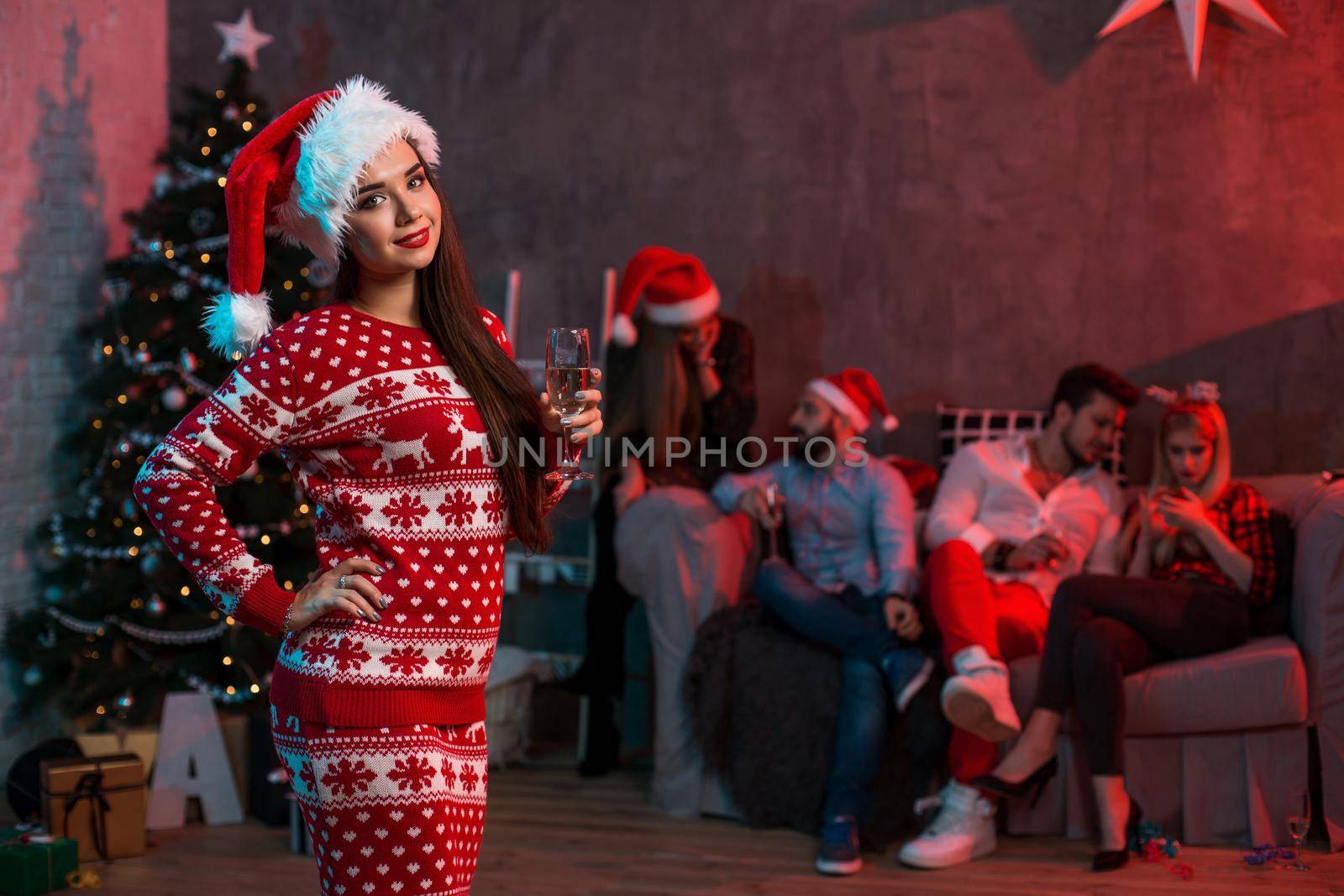 Portrait of a young woman with a glass of champagne on the foreground. Beautiful brunette in a Santa hat, red costume with deers. Christmas party at home.