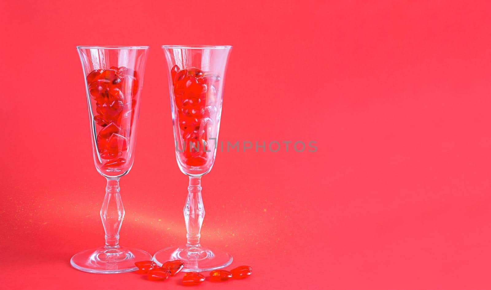 Two champagne glasses are filled with glass hearts on a red background with space for text. Valentine's Day, love, romantic date by Simol