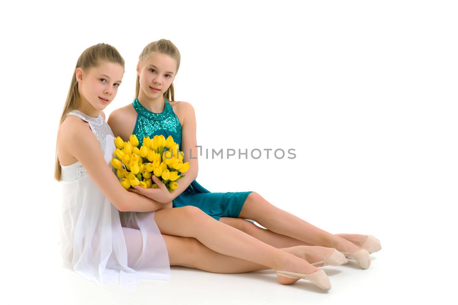 Beautiful girls sitting on the floor with a bouquet of yellow tulips, smiling sisters in summer dresses, portrait of pretty teenage girls on a white background