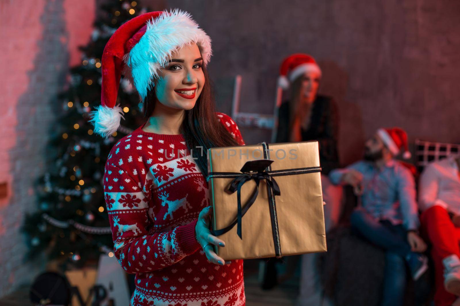 Portrait of a young woman with a gold giftbox at home on the foreground. Beautiful brunette in a Santa hat, red costume with deers by nazarovsergey