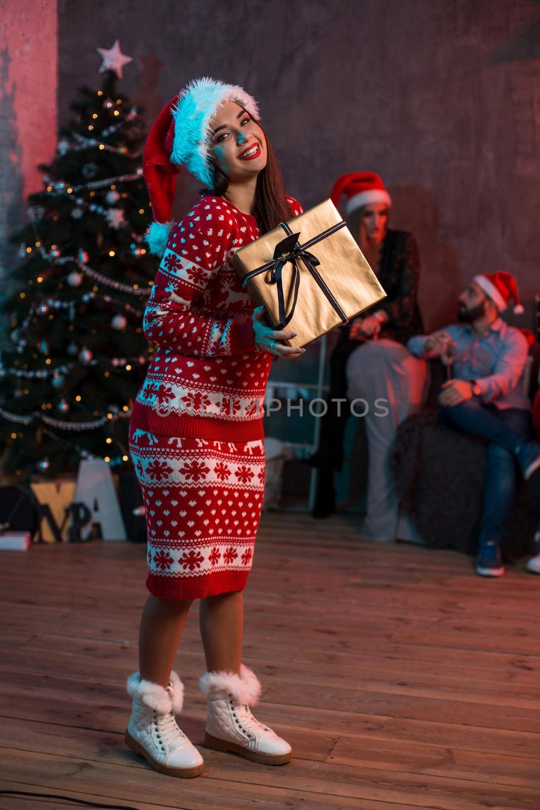 Portrait of a young woman with a gold giftbox on the foreground. Beautiful brunette in a Santa hat, red costume with deers. Christmas party at home.