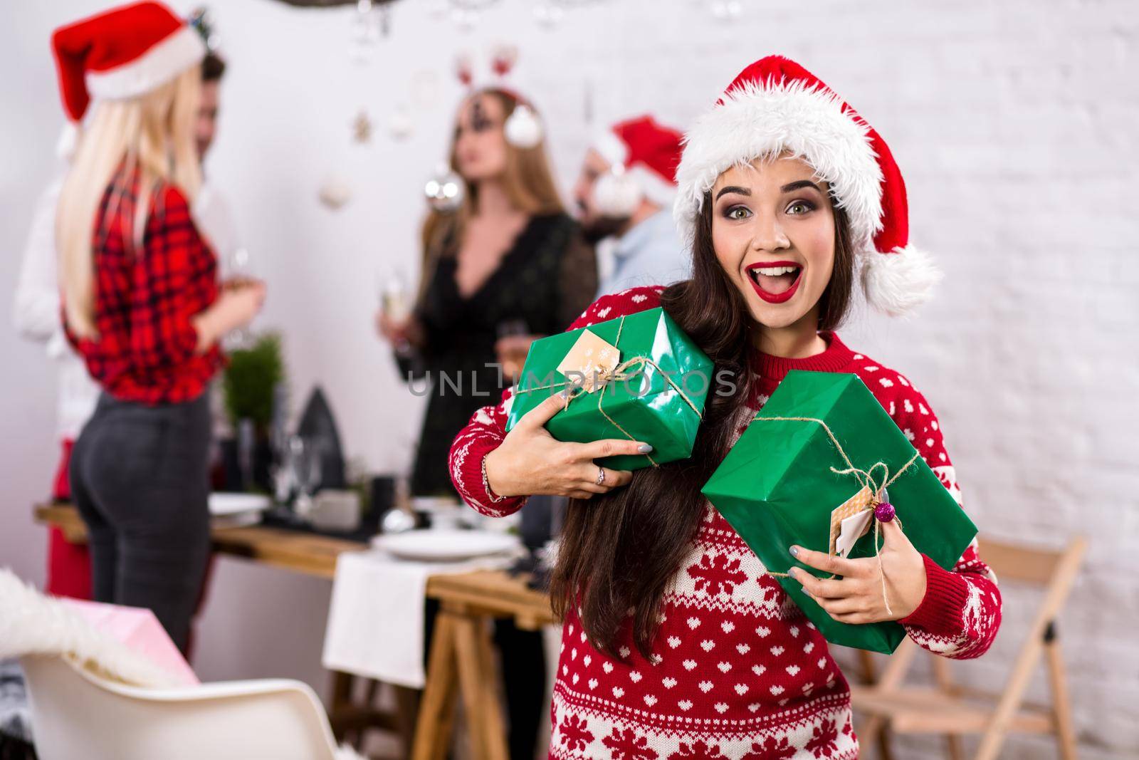 Portrait of a young woman with green giftboxes at home on the foreground. Beautiful brunette in a Santa hat, red costume with deers by nazarovsergey