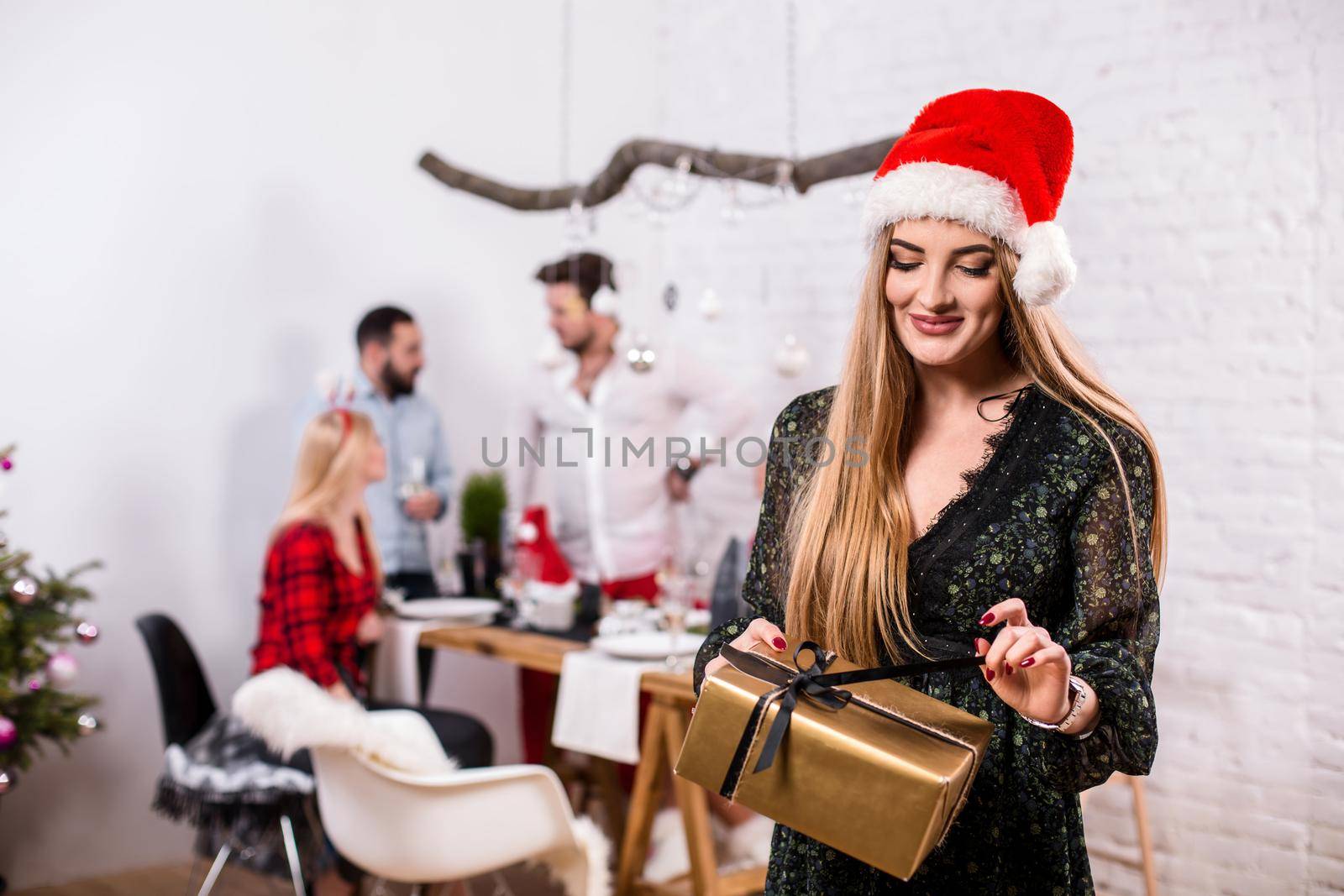 Portrait of a young woman with a gold giftbox at home on the foreground. Beautiful blonde in a Santa hat and black dress by nazarovsergey