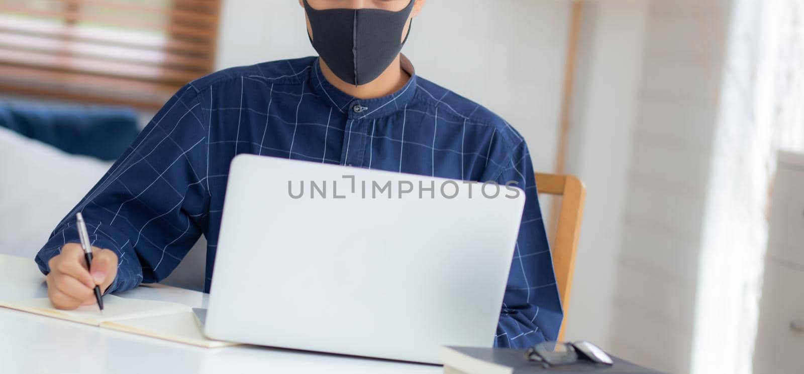 Young asian business man in face mask for protect covid-19 and work on laptop computer and writing notebook, businessman quarantine work from home, new normal, social distancing and stay home. by nnudoo