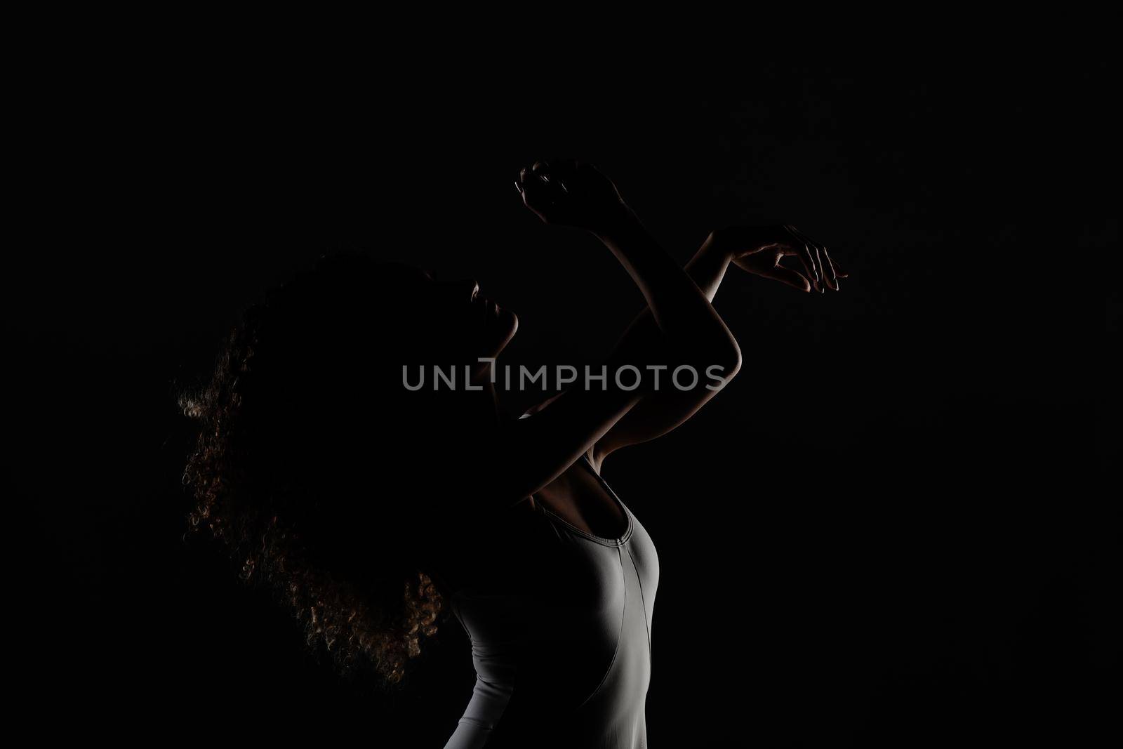Girl with curly hair making ballet poses. Side lit silhouette of ballerina in white dress against black background. by kokimk