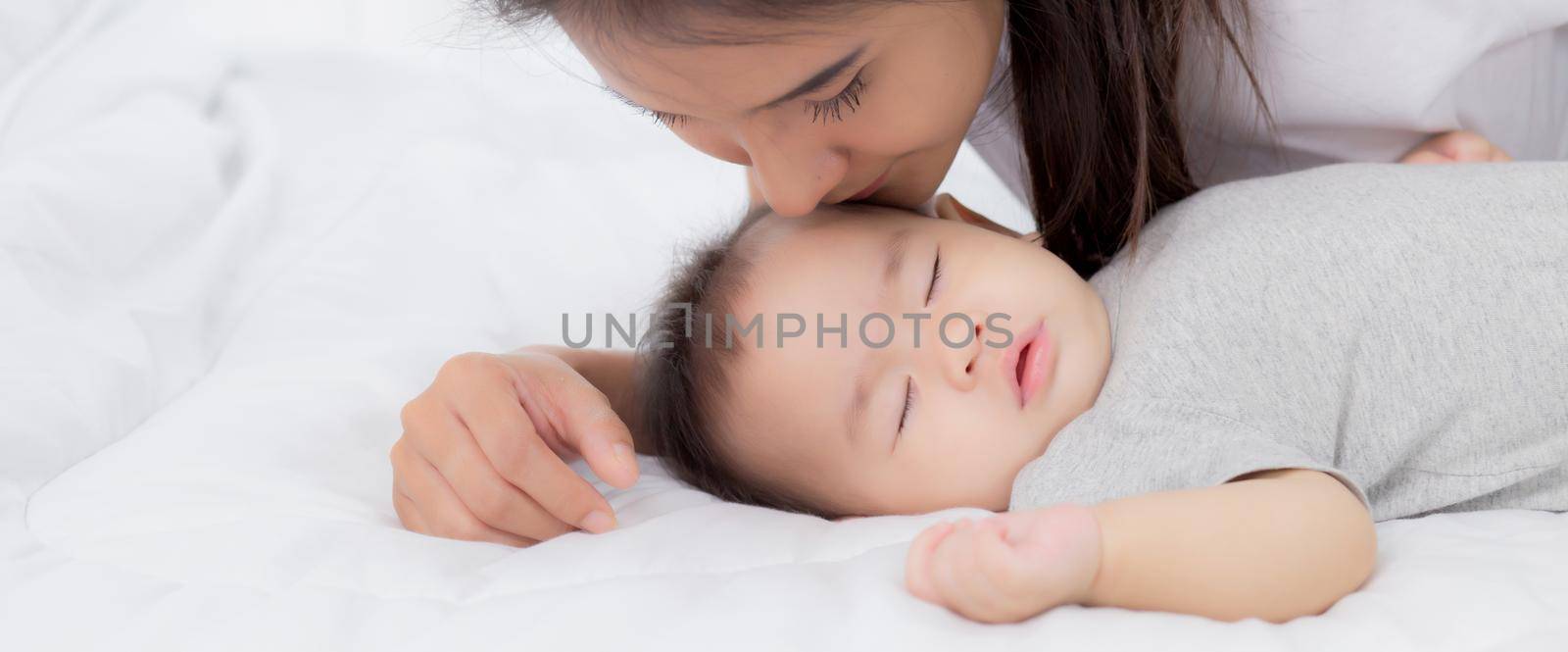 Young asian mother kiss cheek of little baby girl with tender on bed in the bedroom, mom love newborn and care, mother with expression with child together, parent and daughter, family concept.