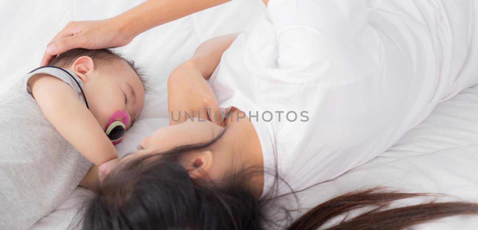 Young asian mother touch head of little baby girl with tender on bed in the bedroom, mom love newborn and care, woman with expression with child together, parent and daughter, family concept. by nnudoo