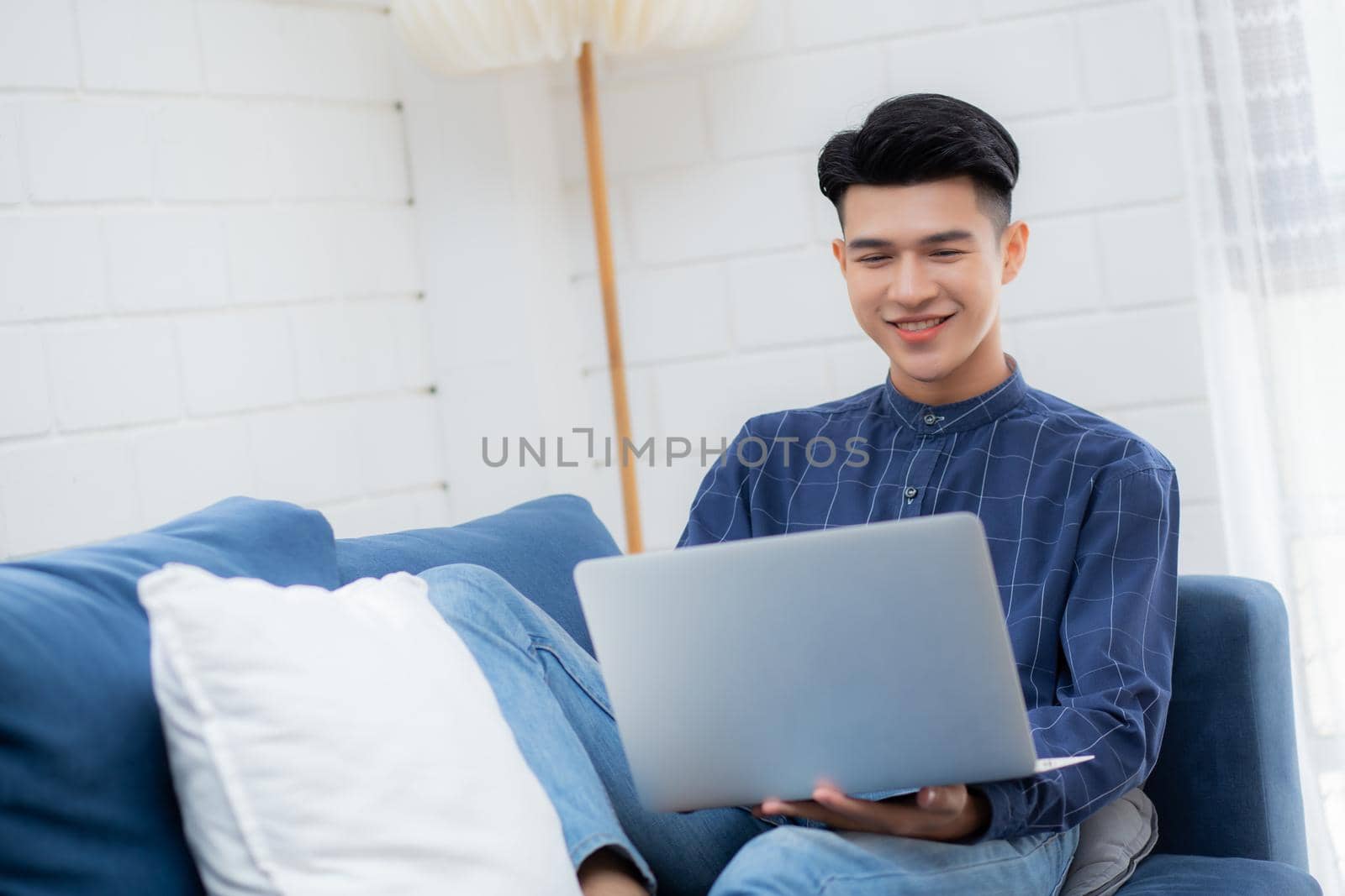 Young asian businessman smile and work from home with laptop computer online to internet on sofa in living room, freelance man using notebook on couch with comfort, new normal, lifestyle concept.