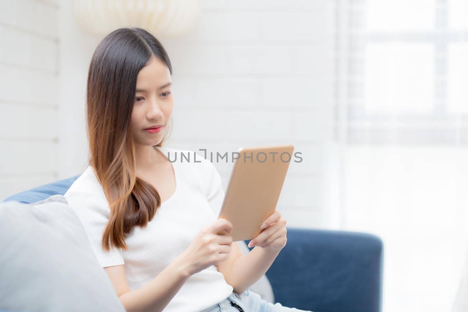 Young attractive asian woman resting using browsing tablet computer on sofa at home, happy girl sitting on couch relax reading digital gadget at house, communication and lifestyle concept. by nnudoo