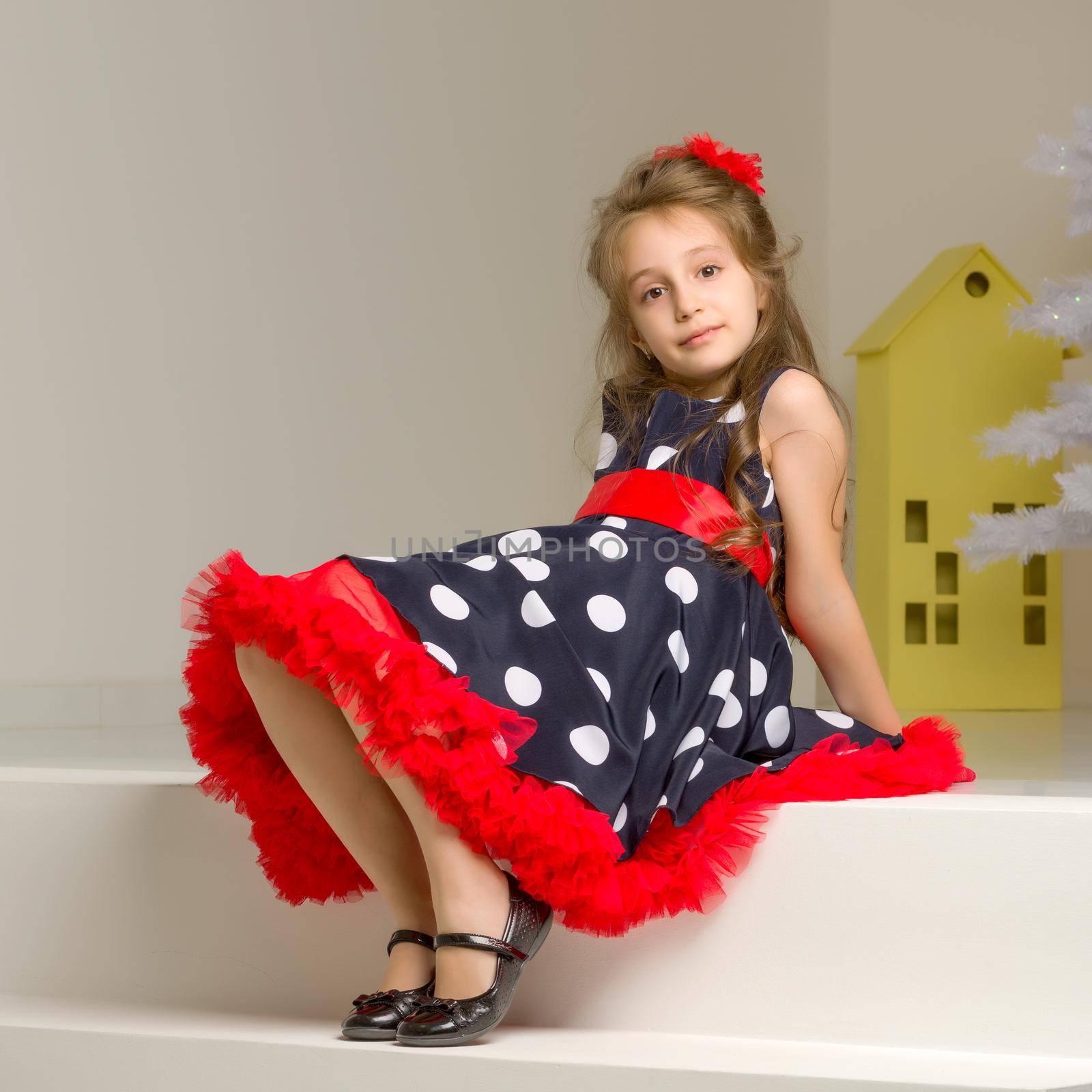 Portrait of Beautiful Girl Wearing Blue Polka Dot Dress, Red Bow and Black Shoes Sitting on White Stairs and Looking at Camera, Charming Coquettish Girl Posing in Retro Fashion Clothes in Studio