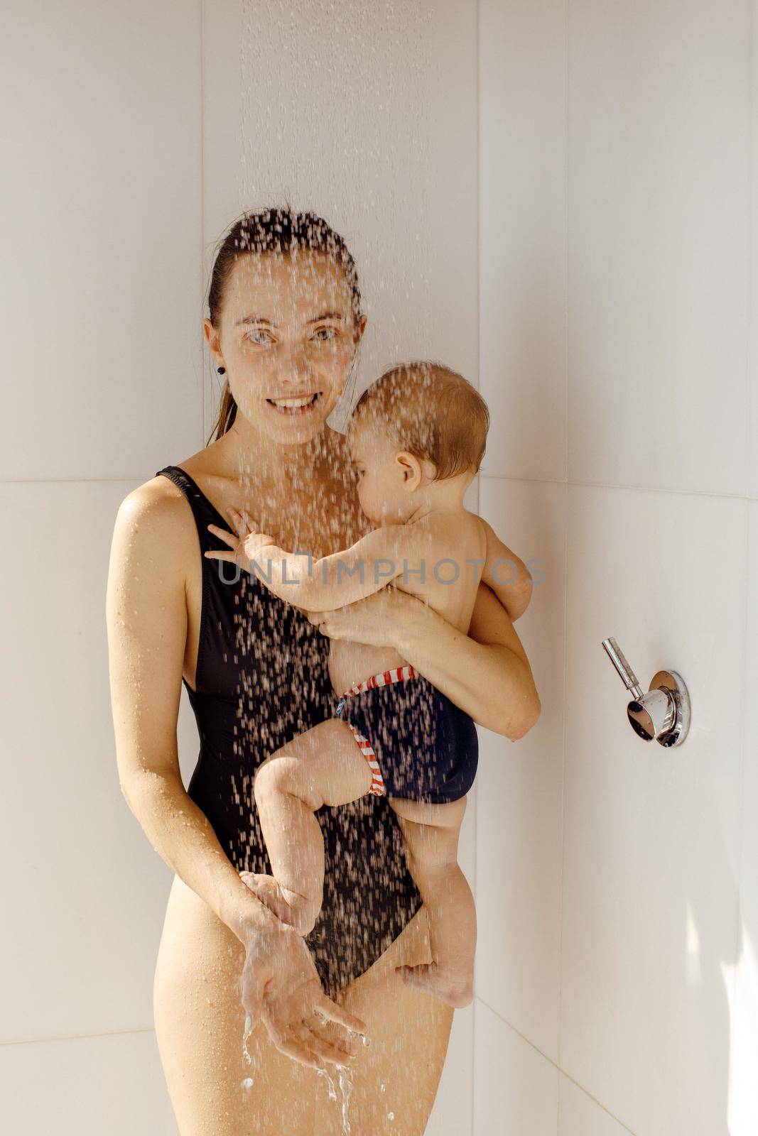 Smiling mother in bathing suit holding on hand baby standing under water in shower and looking at camera
