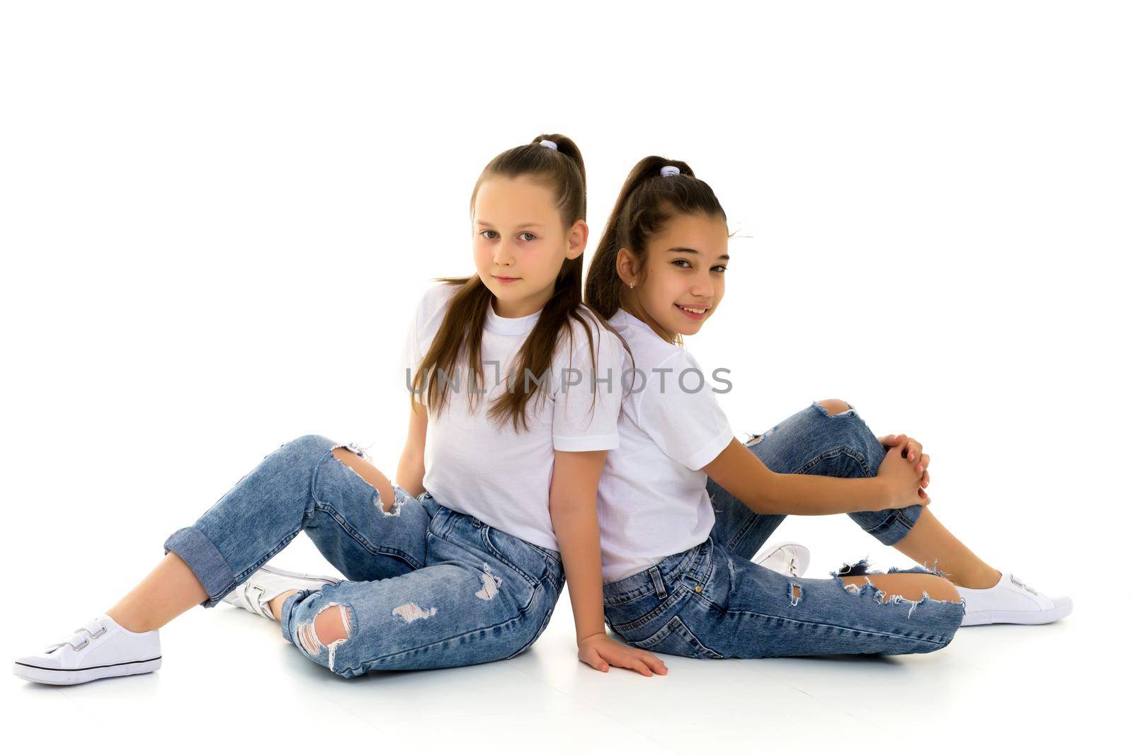 Two cute little girls are sitting on the floor in a studio on a white background. Concepts of style and fashion, happy childhood. Isolated.