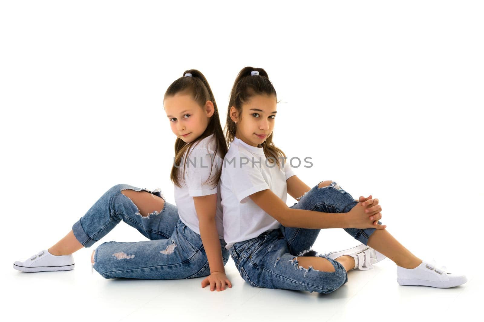 Two cute little girls are sitting on the floor in a studio on a white background. Concepts of style and fashion, happy childhood. Isolated.