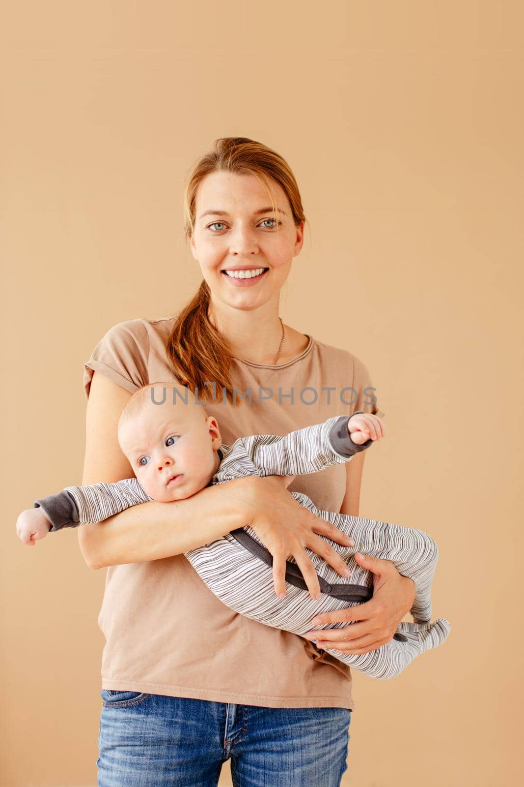 Young attractive mother holding cute adorable baby in hands enjoying and looking at camera on brown background
