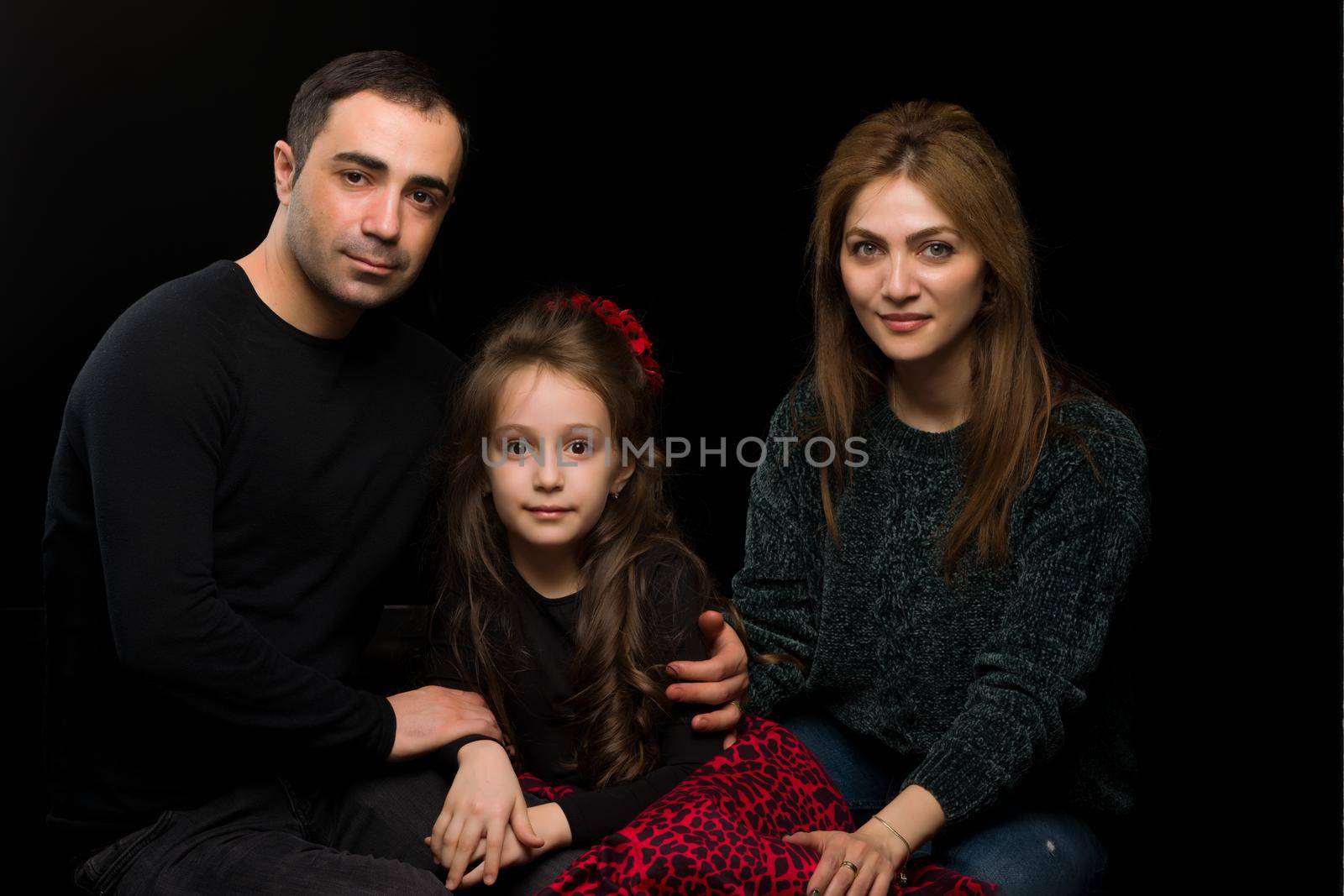 Portrait of Loving Parents Hugging their Adorable Daughter, Mom and Dad Sitting on the Floor and Looking at Their Adorable Girl, Happy Family Sitting Against Black Studio Background
