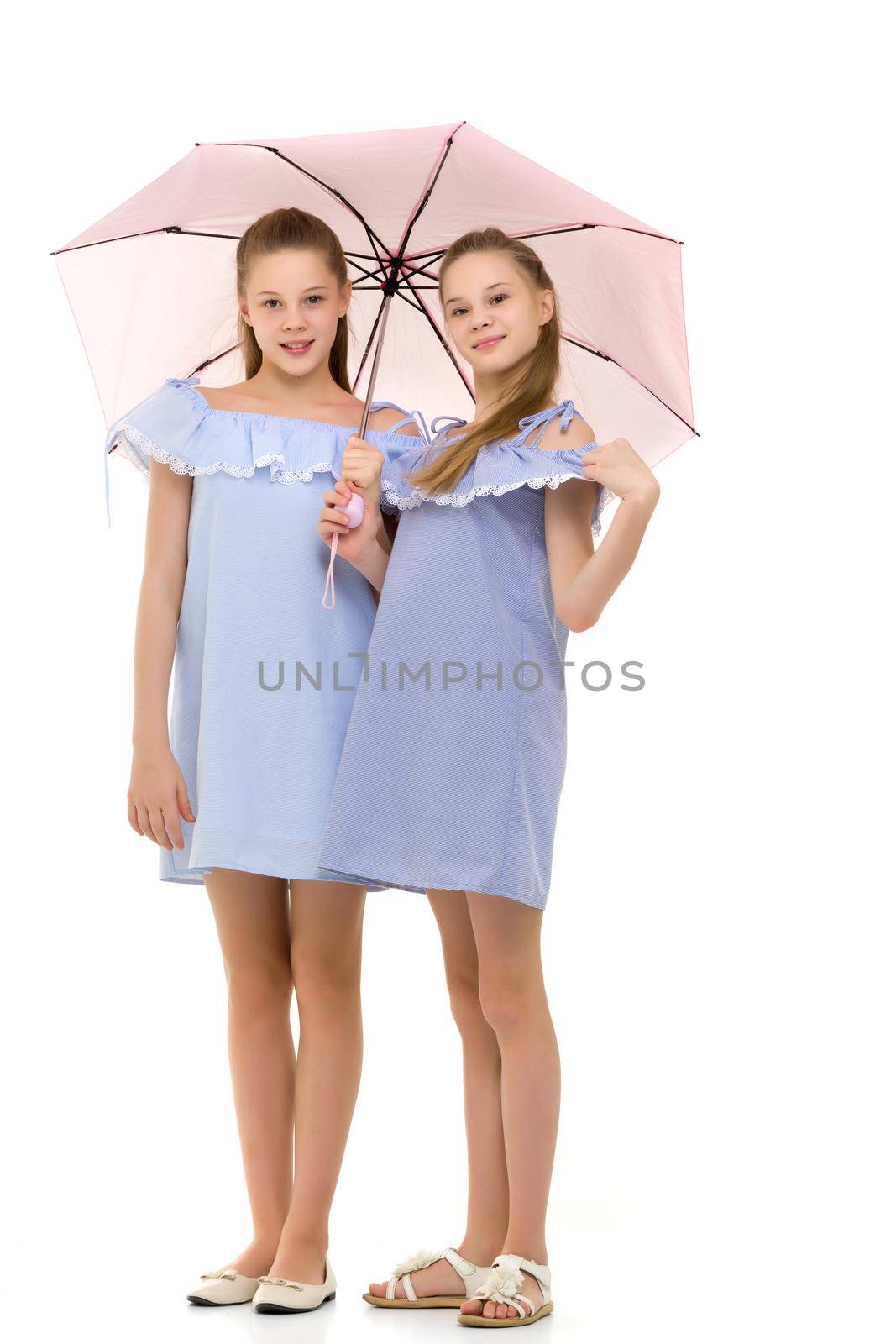 Two Cheerful Pretty Sisters in Identical Light Blue Dresses Standing Under Pink Umbrella, Beautiful Twin Girls in Fashionable Clothes Posing in Studio Against White Background