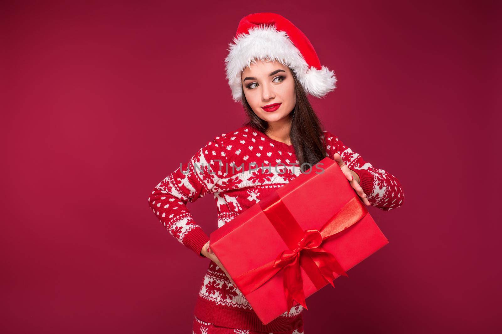 Young woman with long beautiful hair in Santa hat and New Year's dress holding Christmas present in her hands on red studio background. Christmas sale. Emotional brunette. Copy space