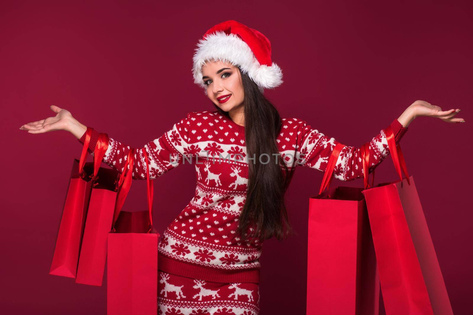 Young pretty woman or girl with long beautiful hair in Santa hat and New Year's dress holding red package on red studio background. Christmas sale. Emotional brunette. Copy space