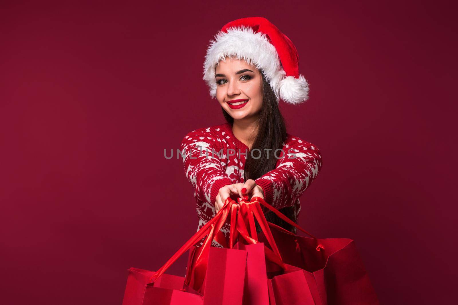 Young pretty woman or girl with long beautiful hair in Santa hat and New Year's dress holding red package on red studio background. Christmas sale. Emotional brunette. Copy space