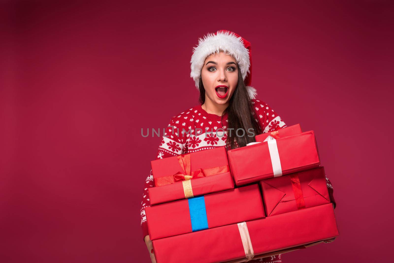 Young woman in sweater and christmas hat holding gift boxes and looking at the camera over red background. Christmas sale. Emotional brunette. Copy space