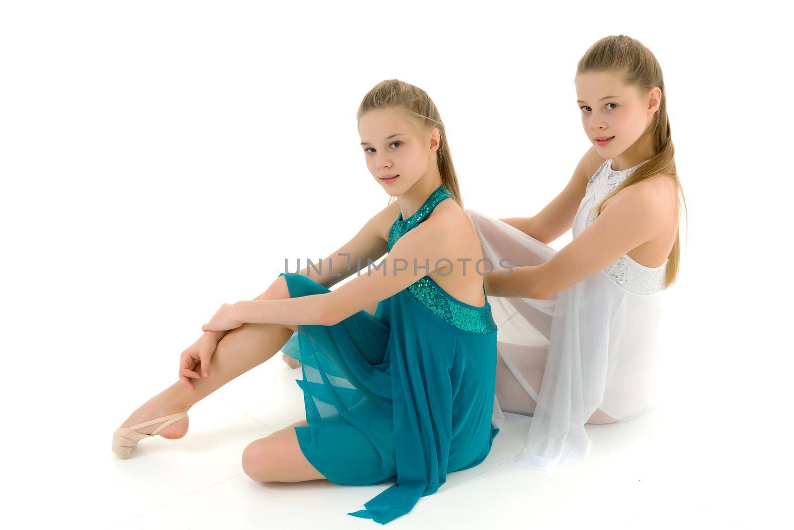 Beautiful Teenage Girls in White and Blue Sport Dresses Sitting on the Floor Against White Background in Studio, Portrait of Two Pretty Twin Sisters Posing Together