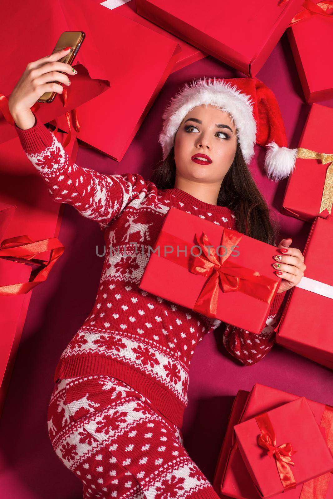 Funny young woman in a New Year's dress lying on a mountain of gifts. Christmas sale. Emotional brunette in Santa hat makes selfie