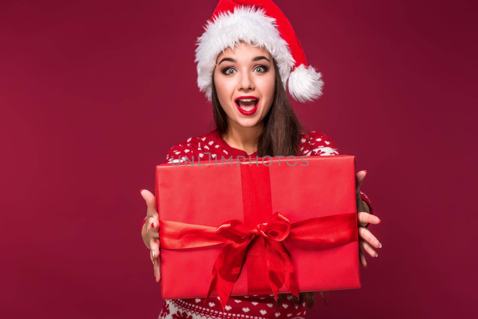 Young woman with long beautiful hair in Santa hat and New Year's dress holding Christmas present in her hands on red studio background. Christmas sale. Emotional brunette. Copy space