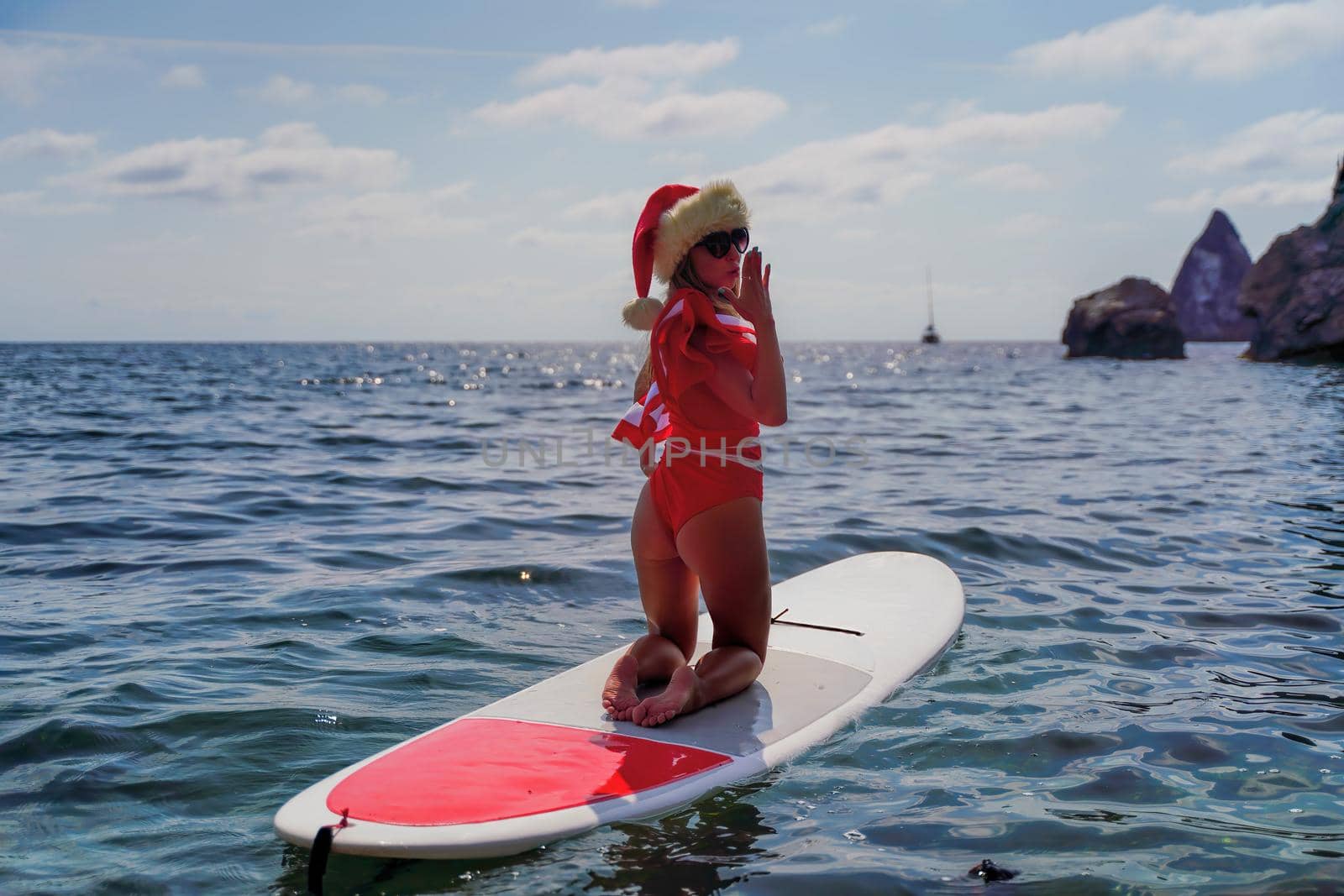 Young brunette woman in red swimsuit and Santa hat, swimming on kayak around basalt rocks like in Iceland. Back view. Christmas and travel concept