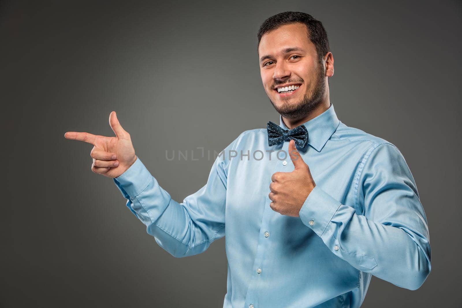 Closeup portrait of excited, energetic, happy, smiling man in blue shirt and butterfly tie is gesturing with his hand, pointing to something with an index finger, isolated on gray background. Positive human emotion facial expression