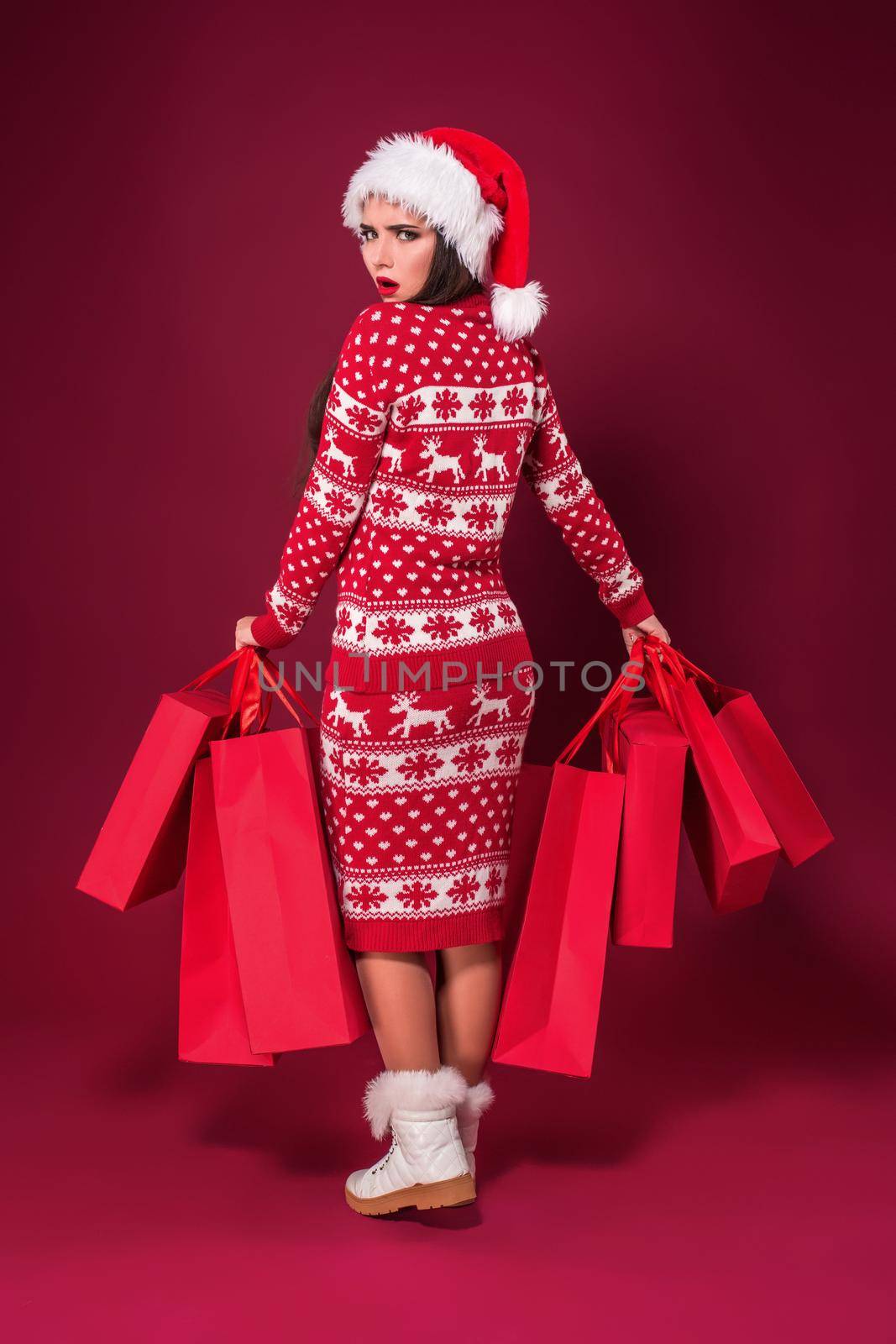 Young pretty woman or girl with long beautiful hair in Santa hat and New Year's dress holding red package on red studio background. Christmas sale. Emotional brunette. Copy space. Full-length photo