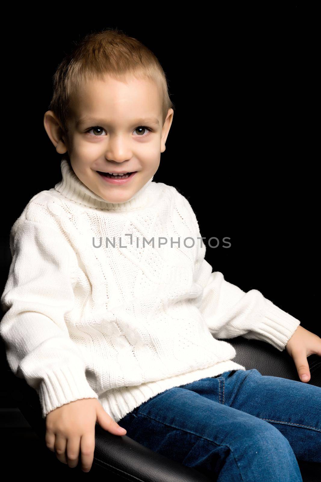 Cute little boy sitting on the armchair in the studio on a black background. The concept of family and people.