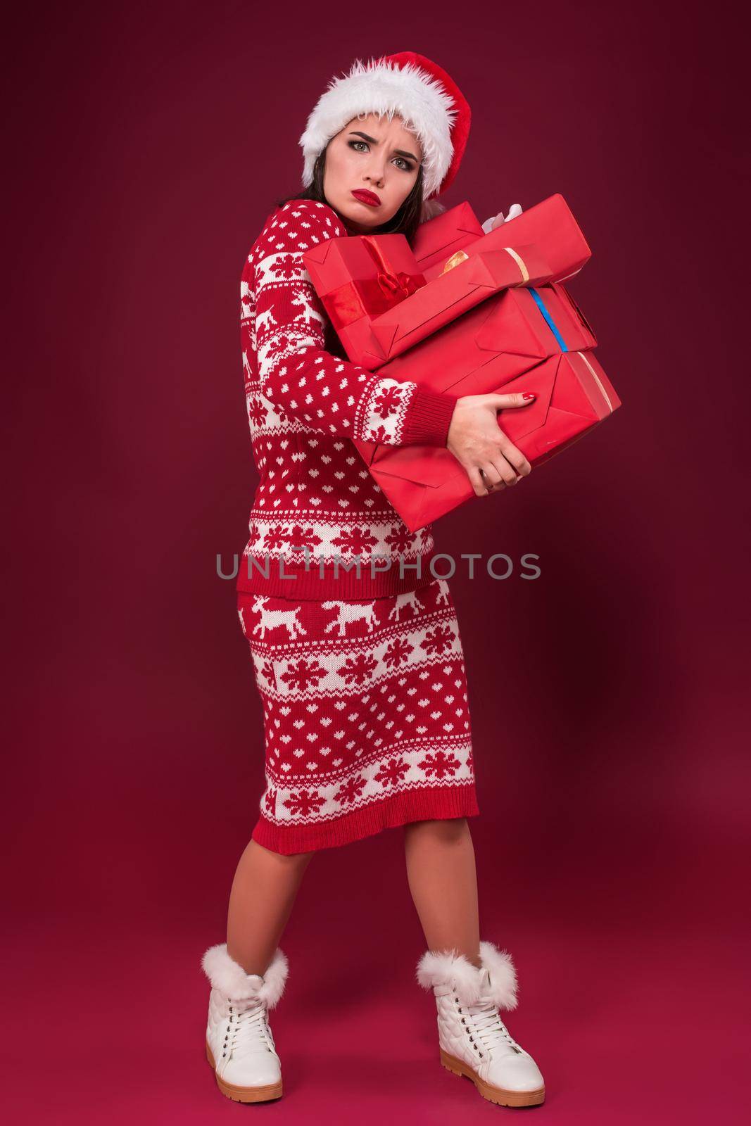 Young woman in dress and christmas hat holding gift boxes and looking at the camera over red background. Christmas sale. Emotional brunette. Copy space