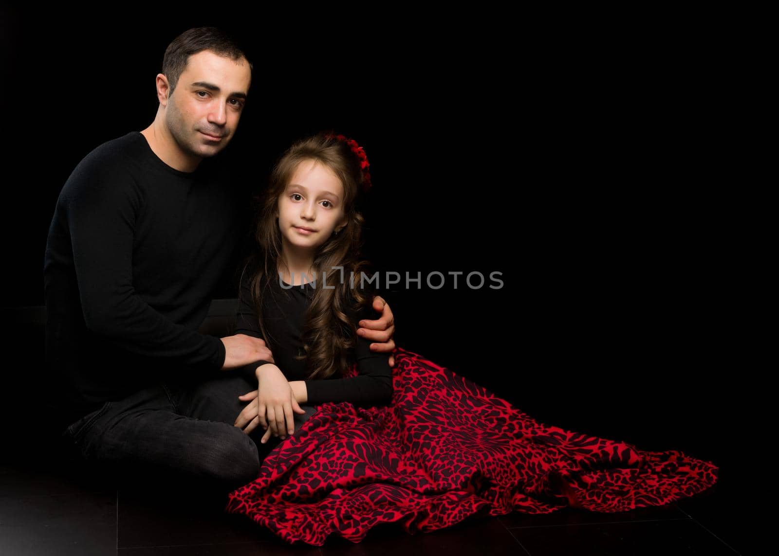 Portrait of Loving Father Hugging His Beautiful Daughter, Handsome Young Man Sitting on the Floor with his Adorable Girl, Happy Family Sitting Against Black Studio Background