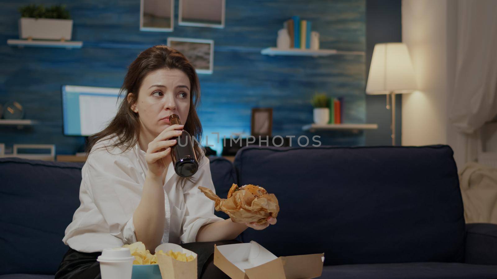 Young person enjoying takeaway burger and beer after work sitting at home on sofa. Woman eating fast food and drinking alcoholic beverage while watching television in living room