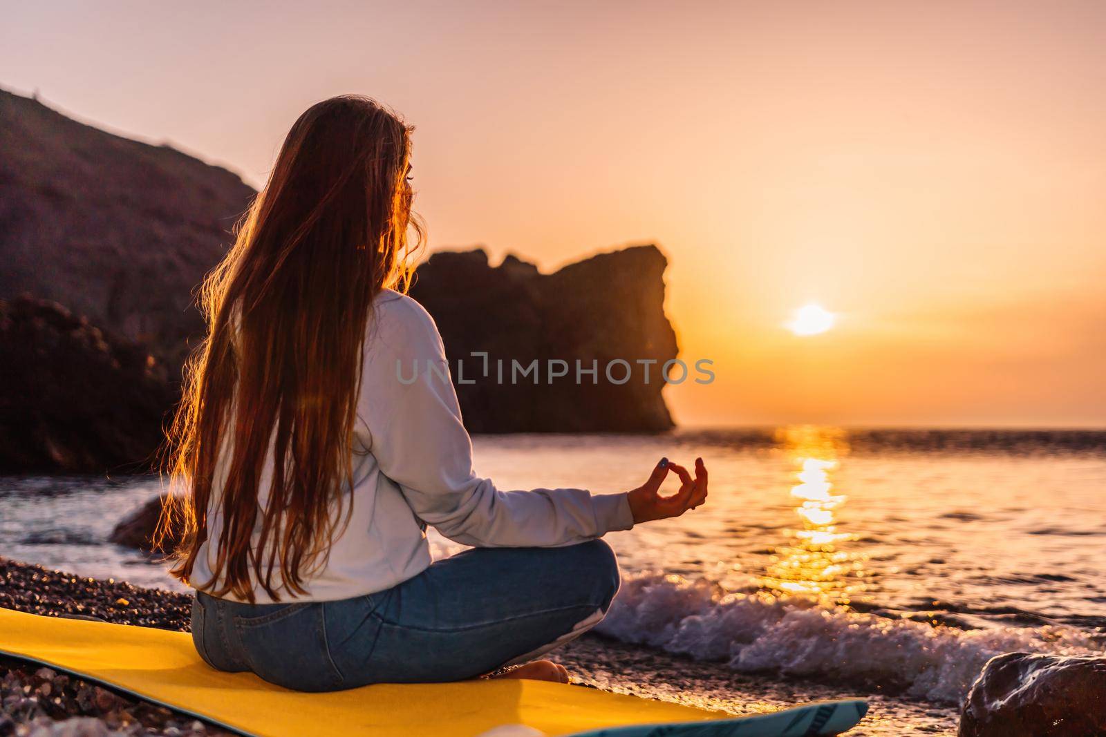 silhoute of Young woman with long hair in blue jeans and sportswear practicing outdoors on yoga mat by the sea on a sunset. Women's yoga fitness routine. Healthy lifestyle, harmony and meditation by panophotograph