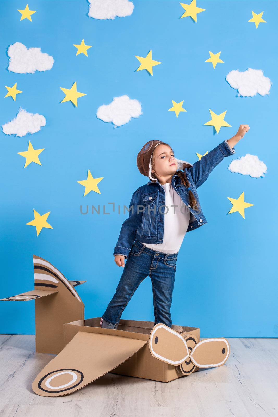 Little child girl in a pilot's costume is playing and dreaming of flying over the clouds. Portrait of funny kid on a background of bright blue wall with yellow stars and white clouds