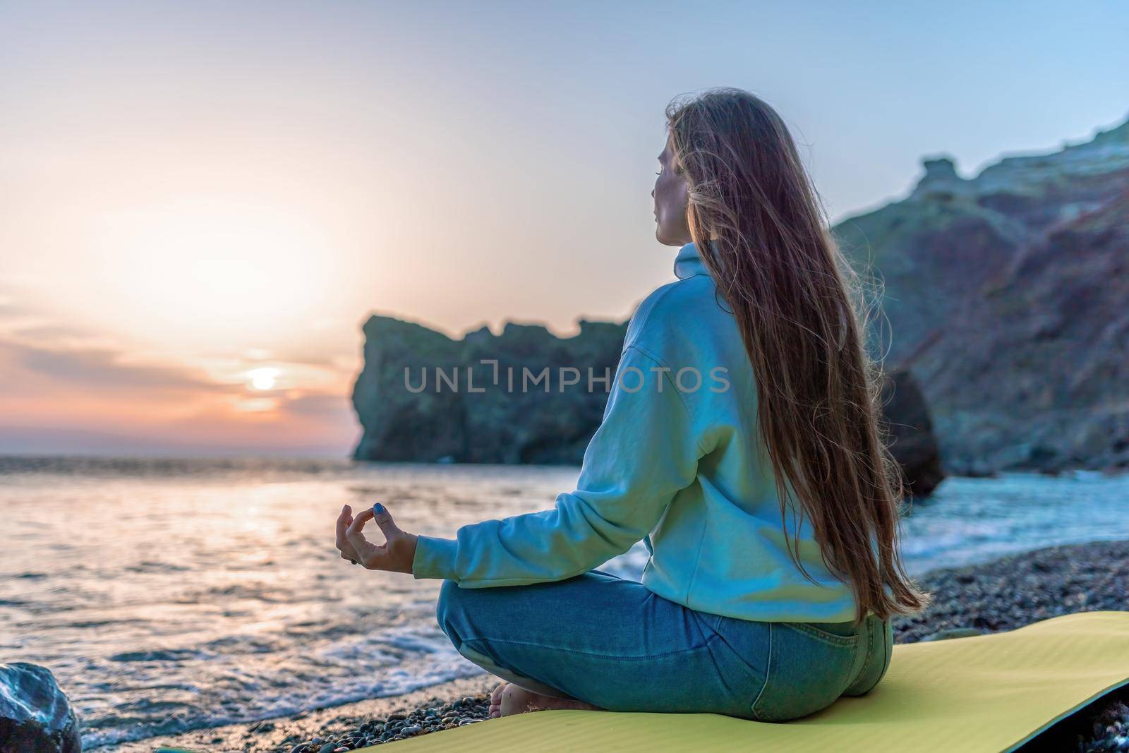 silhoute of Young woman with long hair in blue jeans and sportswear practicing outdoors on yoga mat by the sea on a sunset. Women's yoga fitness routine. Healthy lifestyle, harmony and meditation by panophotograph