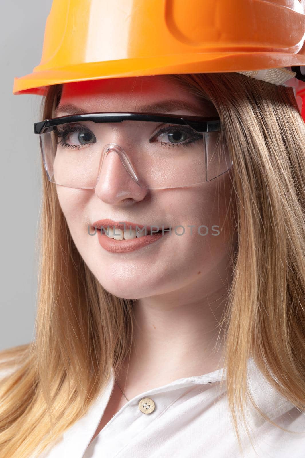 Portrait of a young attractive woman with blond hair in orange helmet and glasses on a neutral gray background.