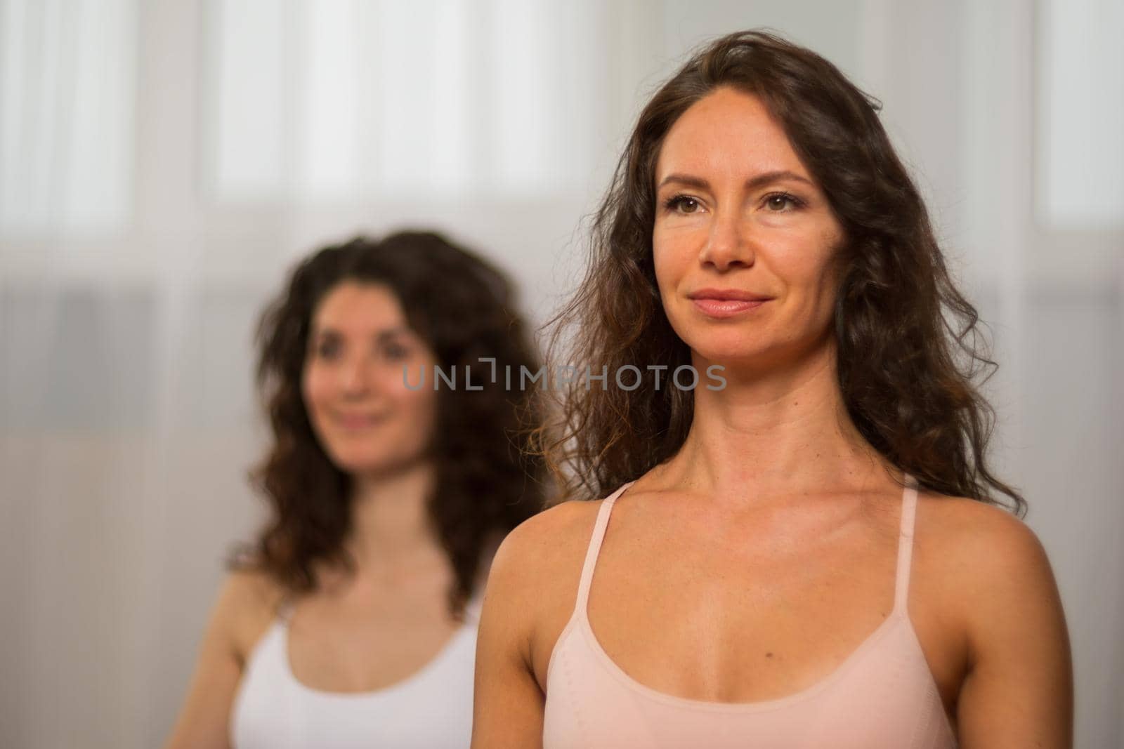 A young woman fitness instructor in violet Sportswear Leggings and Top stretching in the gym before her pilates, on a yoga mat near the large window on a sunny day, female fitness yoga routine concept by panophotograph