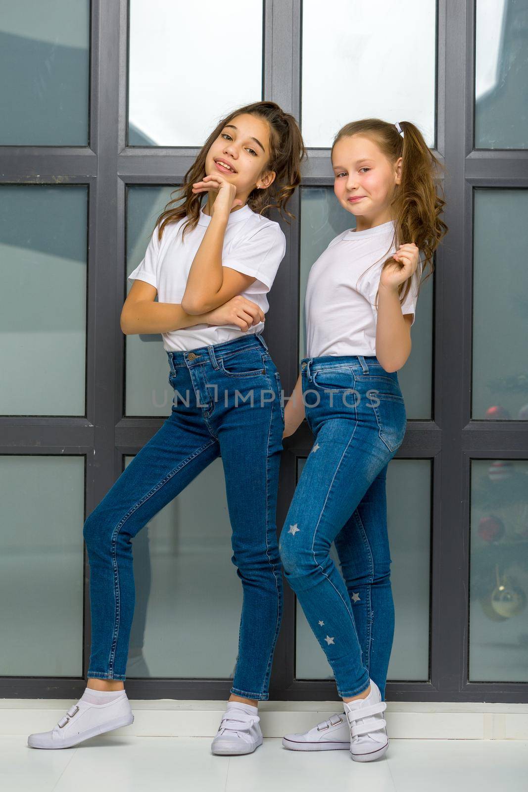 Two beautiful little girls of school age posing in the studio near a large panoramic window. Outside the window it is already evening, dark, the concept of tenderness and beauty.