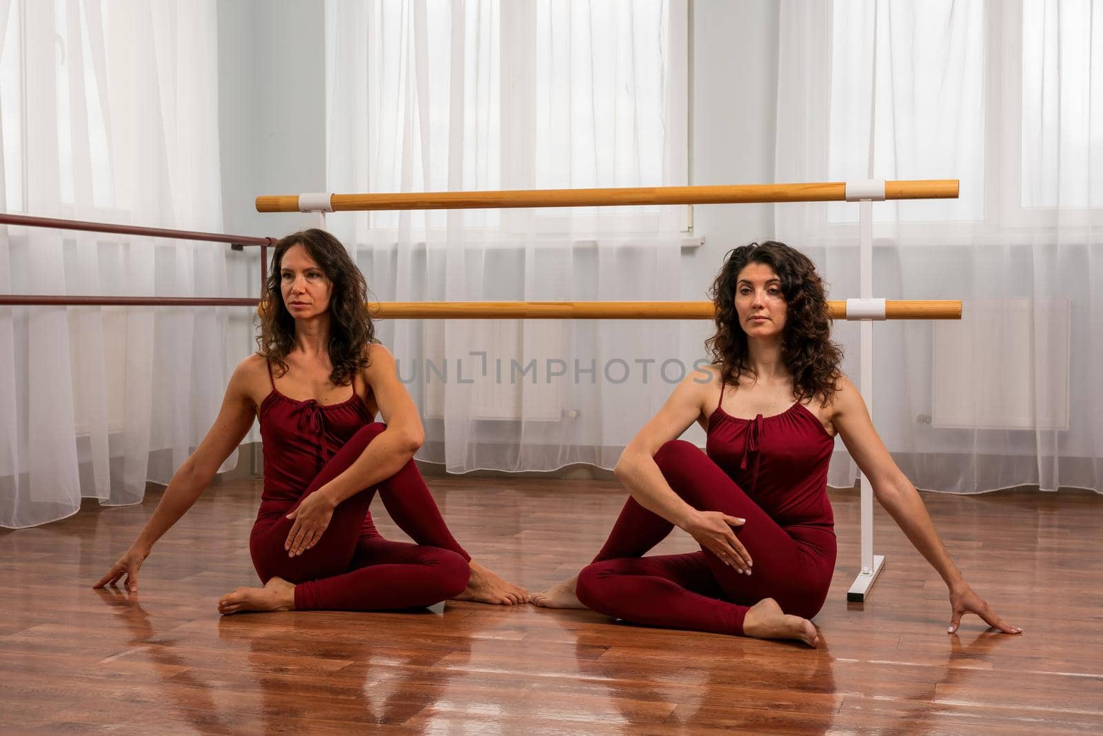 Two young womans fitness instructor in red Sportswear Leggings and Top stretching in the gym before pilates, on a yoga mat near the large window on a sunny day, female fitness yoga routine concept by panophotograph