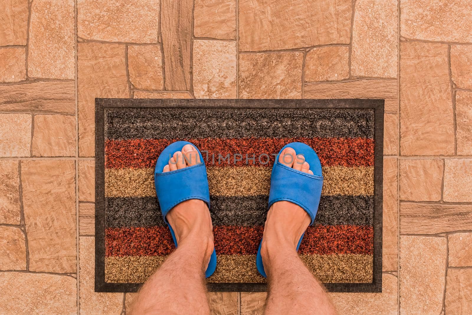 Male feet in blue house slippers stand on a foot mat on a brown tiled floor texture background, top view by AYDO8