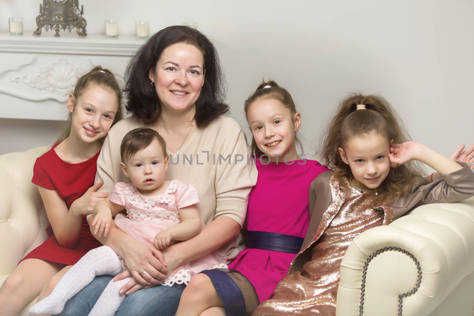 Studio Portrait of Happy Family of Mother and Three Daughters, Smiling Mom and Cute Three Sisters Sitting on the Sofa and Looking at the Camera, Happy Parenting and Childhood Concept