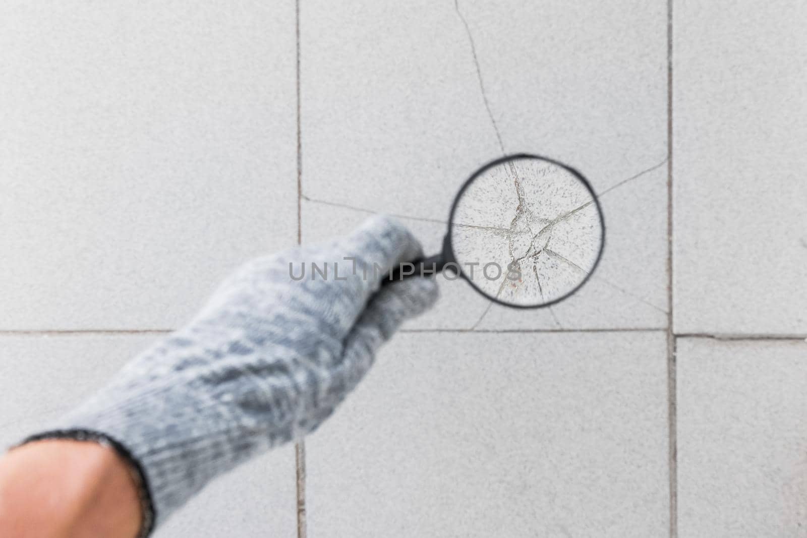 The hand of an industrial worker in a construction glove with a magnifying glass examines the damaged cracked gray tile background. Renovation concept.