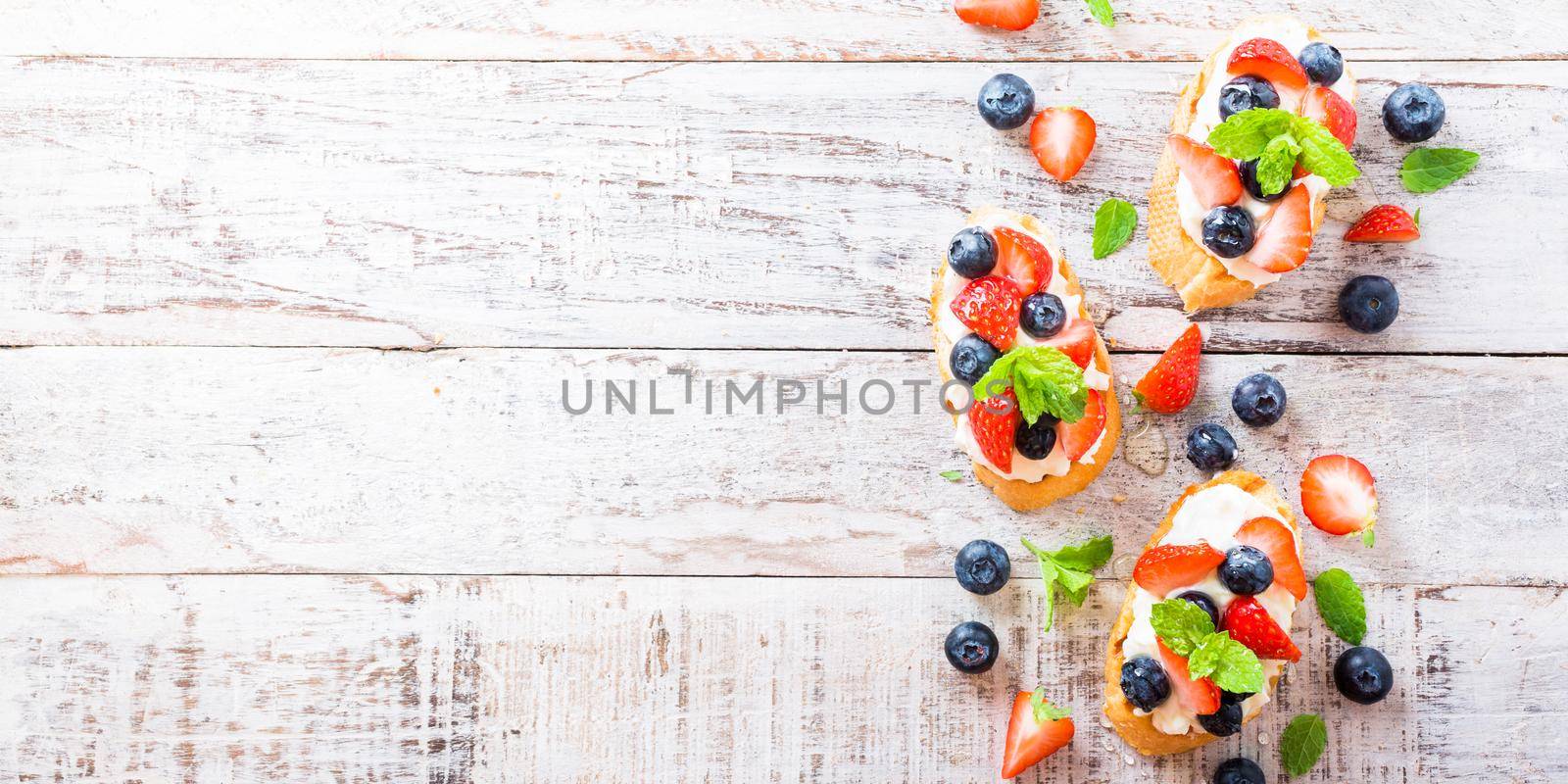 Background with crostini with grilled baguette, cream cheese, blueberry, strawberry, honey and mint on old white wooden background. Delicious appetizer or dessert. Copy space.