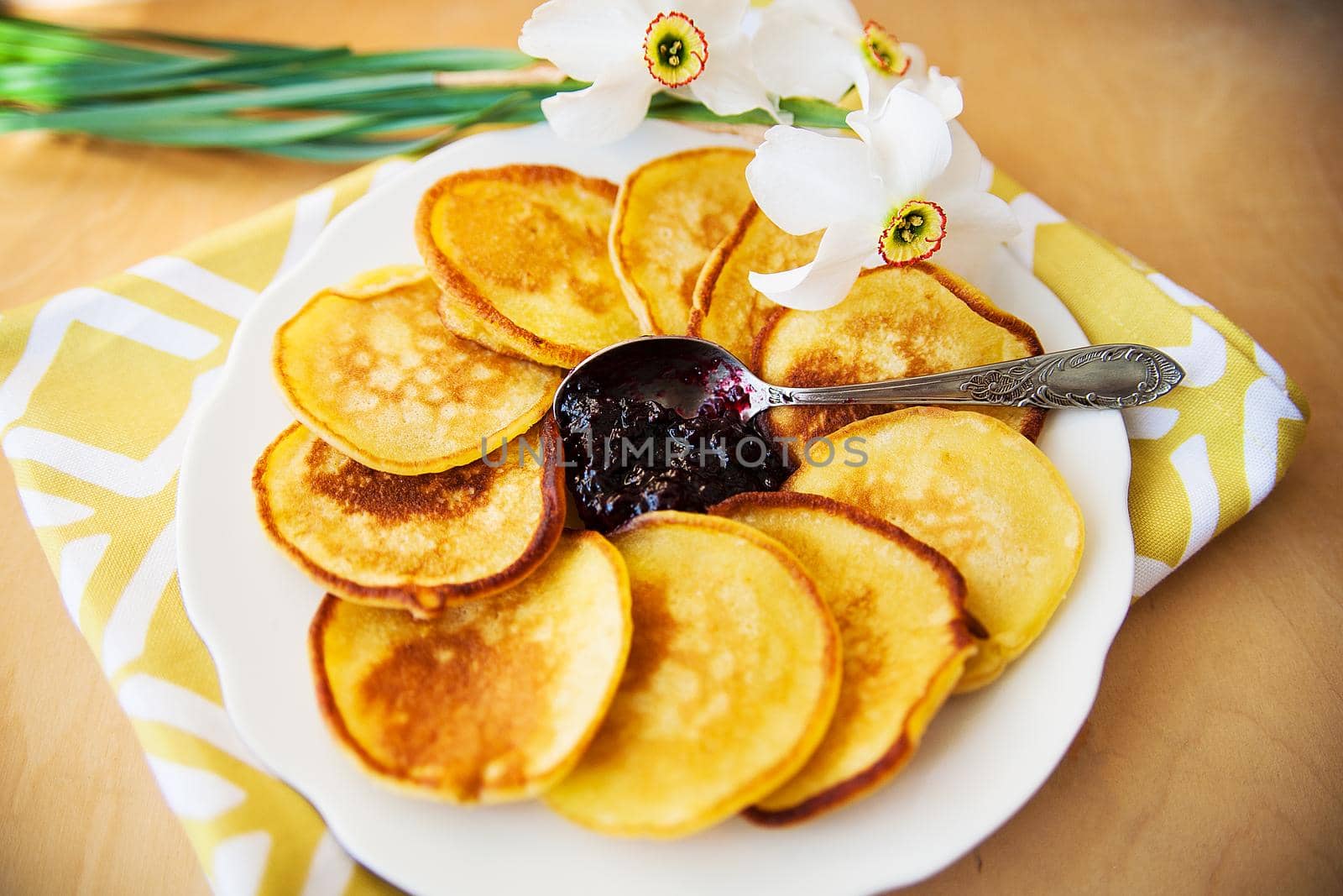 Pancakes with jam and spoon on a plate and white daffodils on wooden background. Top view