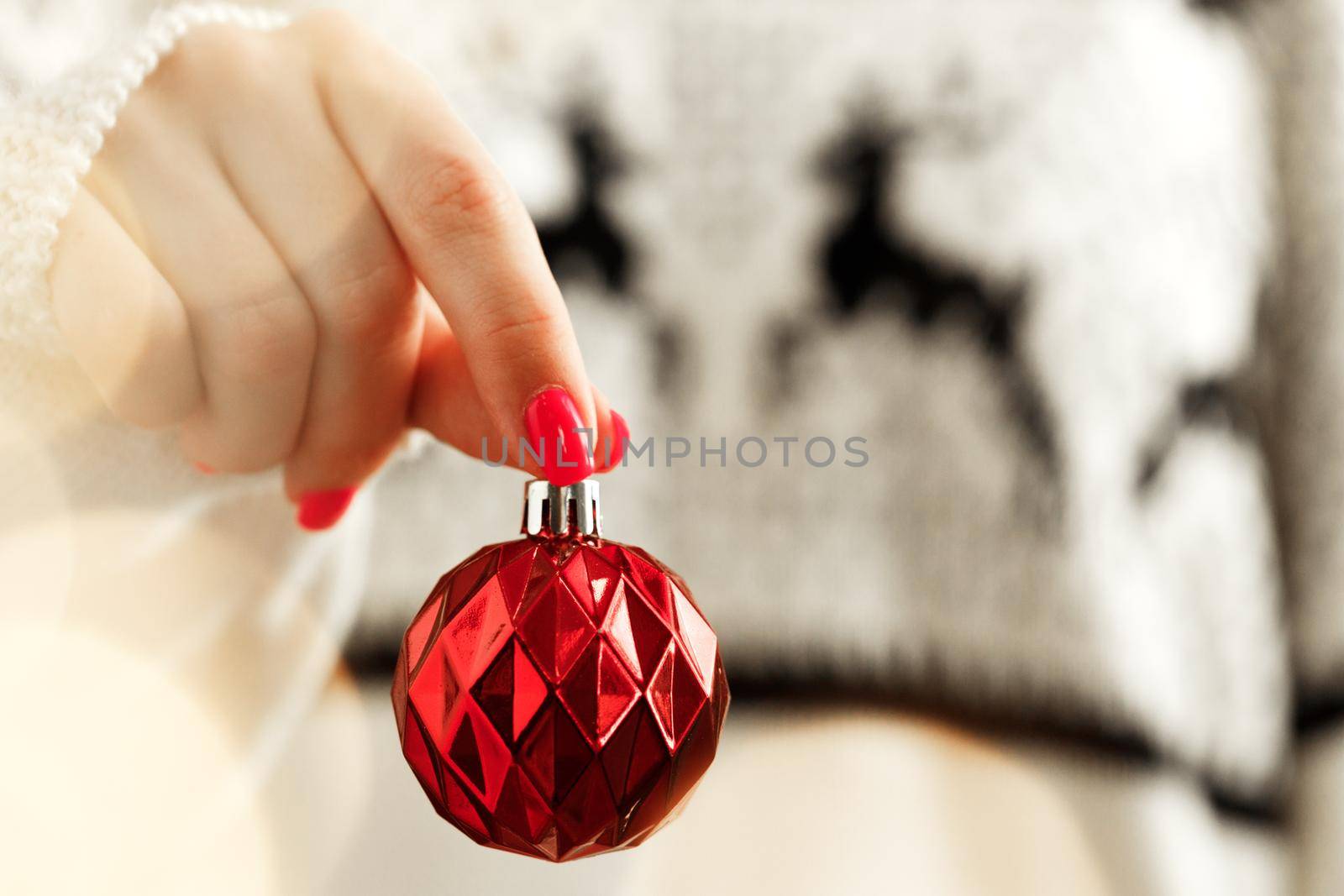 Hand of a woman in festive sweater holding Christmas bauble with blurred lights close up