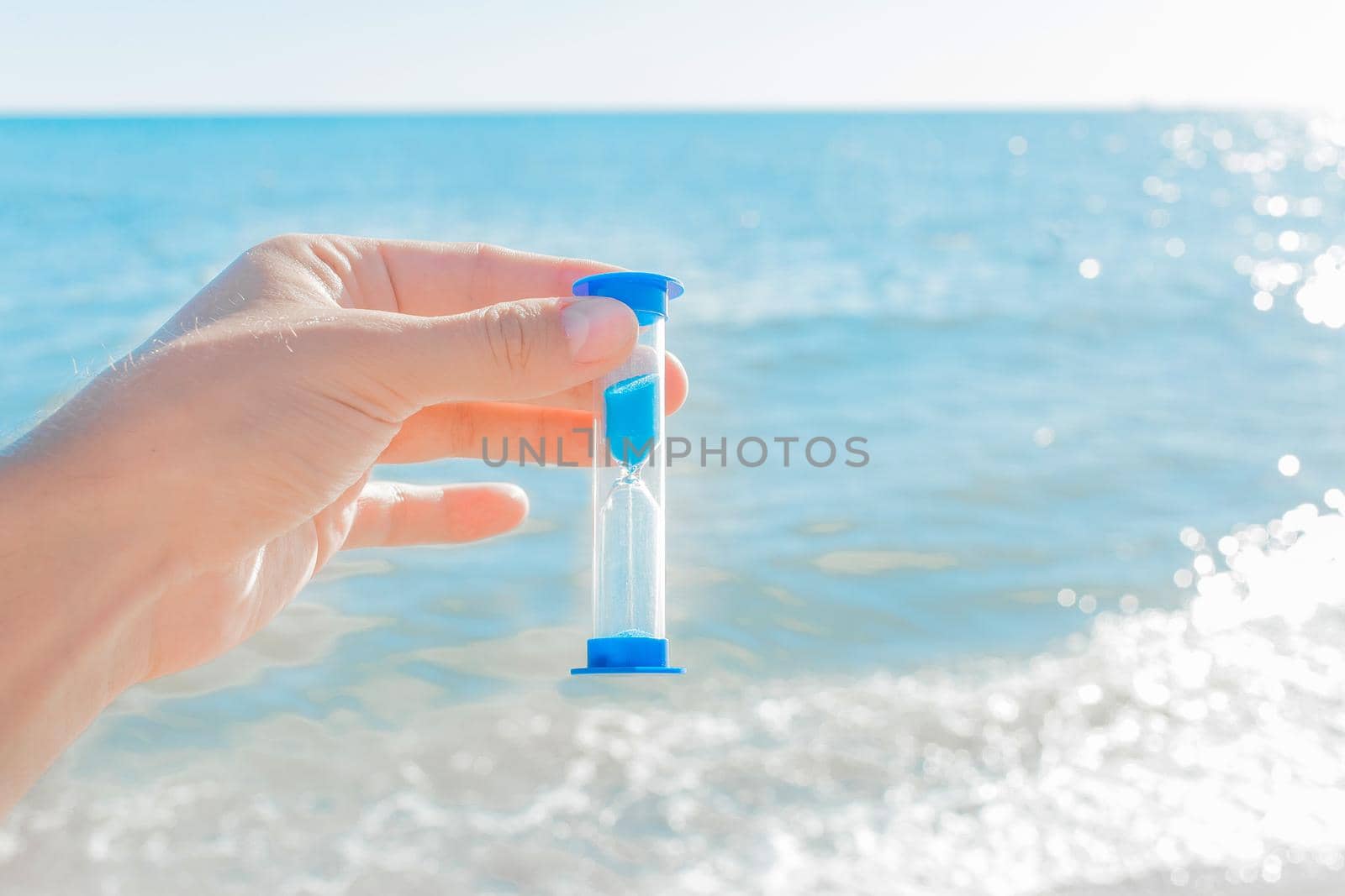 The hand holds the hourglass against the blue bright sea and the sky with a horizon line, close-up.